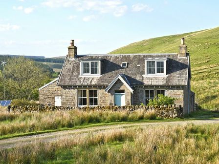 A stone cottage with a tiled roof and chimney in a grassy rural landscape at Roughley Cottage in Newcastleton