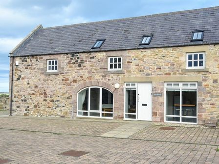 A stone building with multiple windows and a white door on a paved area at Sea Breeze in Newton-by-the-Sea