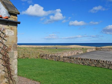 A stone building next to a green lawn with a stone wall field and sea in the background at Snook Point Cottage in Newton-by-the-Sea