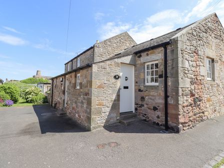 A stone cottage with a white door and windows with a garden and a tower in the background at Wynding Down in Bamburgh