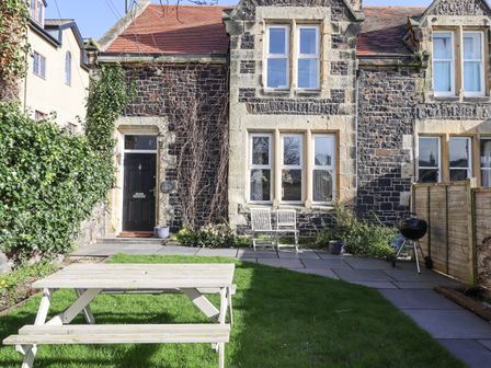 An outdoor garden area with a wooden picnic table on grass in front of a stone house with a black door and white windows at The Hollies in Bamburgh