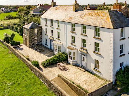 A large white multi-story house with multiple windows and chimneys with a paved patio and garden at No6 Sea Lane in Embleton