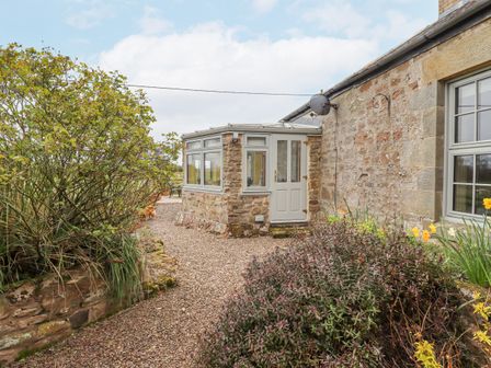 An exterior view of a stone building with a conservatory and gravel path surrounded by shrubs at Adderstone Cottage in Bamburgh
