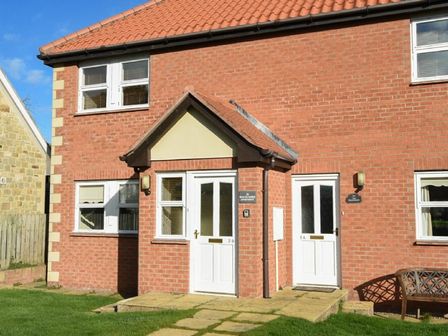 A brick building with white doors and windows and a bench on grass at Beachcomber Apartment in Bamburgh