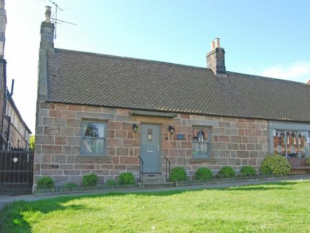 A stone cottage with a tiled roof green door and windows with shrubs in front at Charlton Cottage in Bamburgh