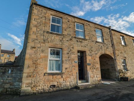 The exterior of a stone building with windows a black door and an arched passage at Riverside Cottage in Warkworth
