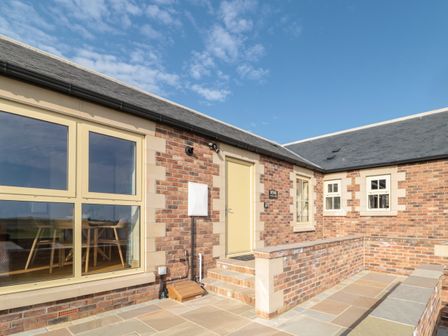 An exterior view of a brick building with beige framed windows and doors and a stone tiled patio at Byre Cottage in Embleton