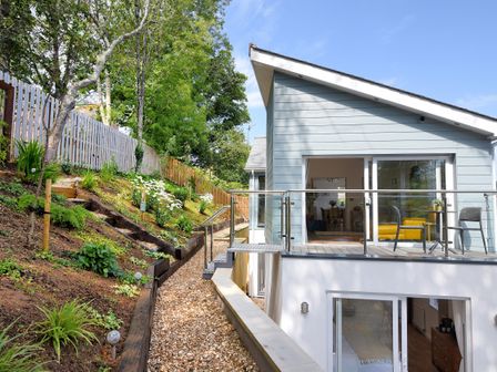 An outdoor area with gravel path flowers and trees next to a modern house with balcony and glass railing at Blue Neptune in Lyme Regis