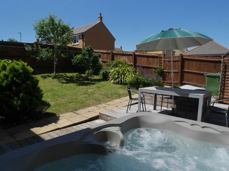 A backyard with a hot tub, a table with chairs, an umbrella, grass, trees, and a wooden fence at Cliffwalk Cottage in Southwell