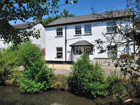 A white two-story house with a porch and multiple windows next to a small body of water with green bushes at River Lym Cottage in Lyme Regis