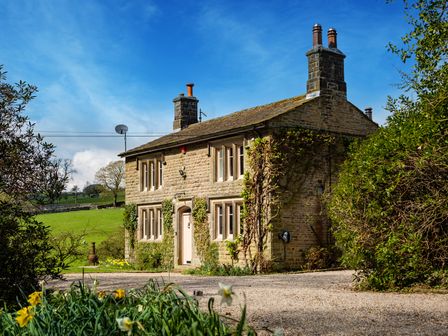 A stone house with a gravel driveway and flowering plants surrounded by trees at The Lodge in Hartlington near Grassington