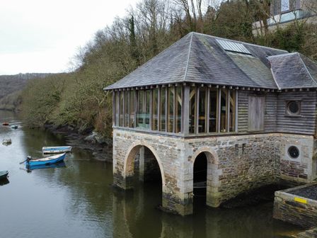 A stone boathouse with large windows on a riverbank with several small boats in the water at Oakenhurst in Golant