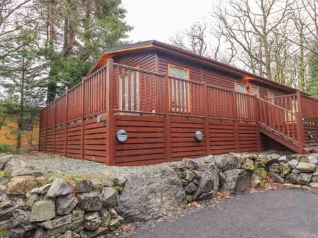 A wooden cabin with a raised deck and stairs surrounded by trees and a stone wall at 25 Thirlmere in Troutbeck Bridge