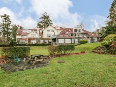 A large building with multiple chimneys surrounded by a grass lawn and bushes with a wooden picnic table and green bench in view at Lake House Loft in Bowness-On-Windermere