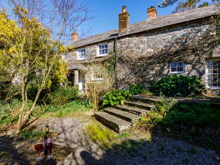 The exterior of a stone house with three chimneys a small porch and stone steps surrounded by plants and trees at Aarons Cottage in St Teath