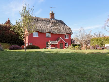 A red house with a thatched roof and a garden with a tree and outdoor seating at Rhubarb Cottage in Ufford near Woodbridge
