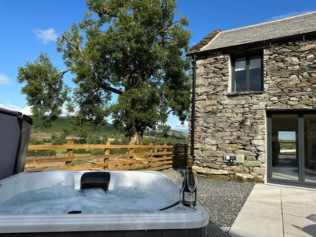 An outdoor jacuzzi with a bottle and two glasses near a stone cottage with a tree and wooden fence at Meadow Syke Byre in Bampton Cumbria