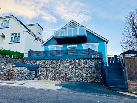 A blue house with a stone retaining wall and a balcony above a street at Ingledene in Brixham