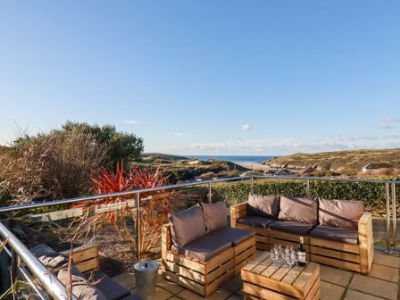 An outdoor patio with wooden sofas and a table holding wine and glasses overlooking a coastal landscape at Seacroft in Crantock