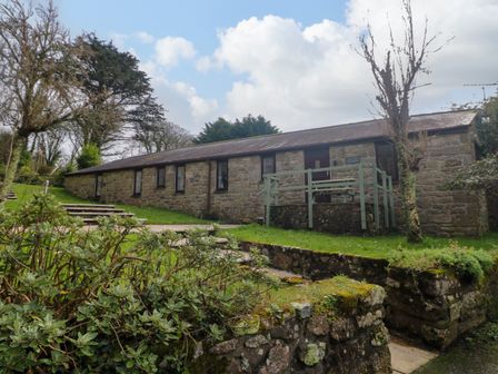 A stone cottage with multiple windows and a small fenced porch surrounded by trees and bushes at The Old Piggeries in Gulval near Penzance