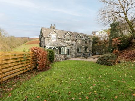 A stone house with a lawn and patio furniture surrounded by trees and a wooden fence at Scandale Bridge Cottage in Ambleside