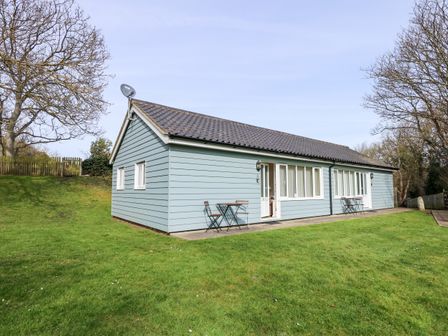 A blue single-story house with two small tables and chairs on a concrete patio surrounded by grass and trees at 11 The Dell in Mundesley