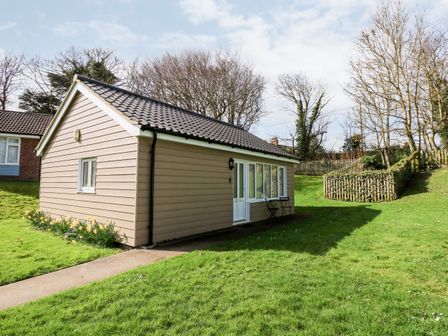 A small beige house with a tiled roof on a grassy lawn with a pathway and wooden fence at 12 The Dell in Mundesley
