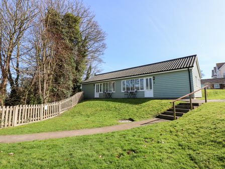A single story house with a tiled roof and green siding on a grassy slope with steps and a wooden fence at 14 The Dell in Mundesley