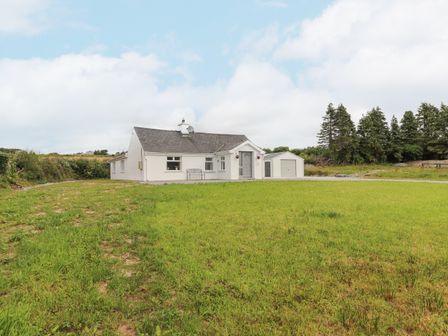 A white single-story house with a gray roof and a small shed in the background surrounded by grass and trees at White Cottage in Abbeyfeale County Limerick