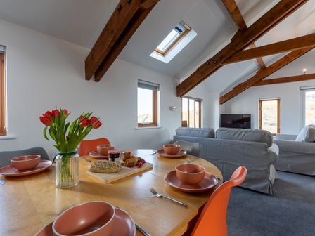A dining area with a wooden table set with dishes and a vase of flowers next to a living room with sofas and wooden ceiling beams at Sunset View in Widemouth Bay
