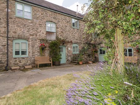 A stone exterior of a house with green framed windows and doors with benches and flowers in front of Blackberry Cottage in Slapton