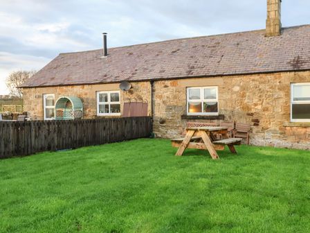 A stone cottage with a wooden fence and outdoor wooden picnic table on a grass lawn at Garden Cottage in Alnmouth