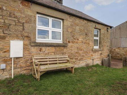An outdoor stone wall with two windows and a wooden bench on grass at Pond Cottage in Alnmouth