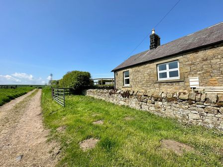 A stone cottage with a slate roof beside a dirt path and grassy area at Lonnon Cottage in Alnmouth