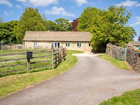 A stone cottage with a tiled roof along a paved driveway with wooden fences and trees at Stonewell Cottage in Stow-On-The-Wold