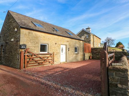 A stone house with a white door and two windows with a wooden gate and gravel driveway at Stable Cottage in Hallbankgate