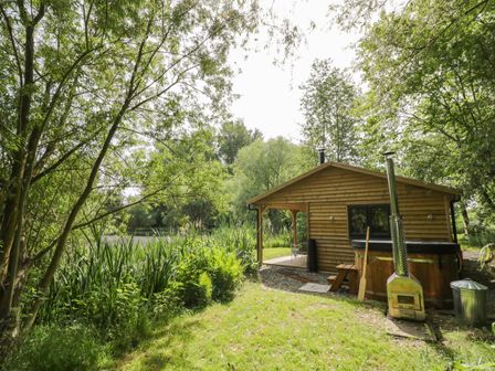 A wooden cabin with a covered porch and outdoor wood stove next to a grassy area and trees at Kingfisher in Dunchurch