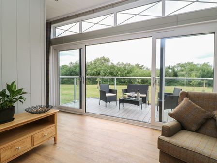 A living room with a wooden cabinet plant and armchair next to sliding glass doors leading to a balcony with outdoor furniture and a grassy area at Holly Lodge in Winthorpe near Newark-On-Trent