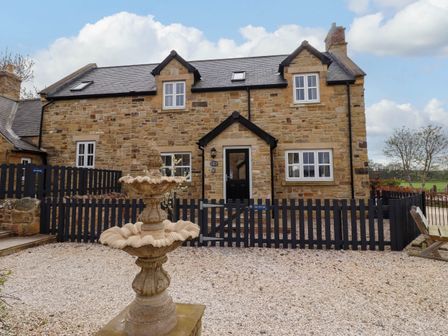 A stone cottage with a black door black fence and a stone fountain in the front garden at Oak Cottage in Lucker near Warenford