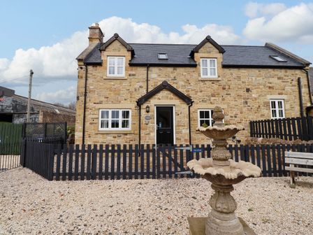 A stone cottage with black door and windows and a two tier stone fountain in gravel yard at Yew Cottage in Lucker near Warenford