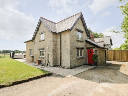 A stone house with a red door and multiple windows surrounded by grass and a gravel driveway at 2 Dewars Farm Cottages in Middleton Stoney