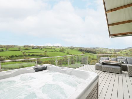 An outdoor deck with a hot tub glass railing and outdoor seating overlooking green hills at Barcud Coch - Red Kite in Garthmyl