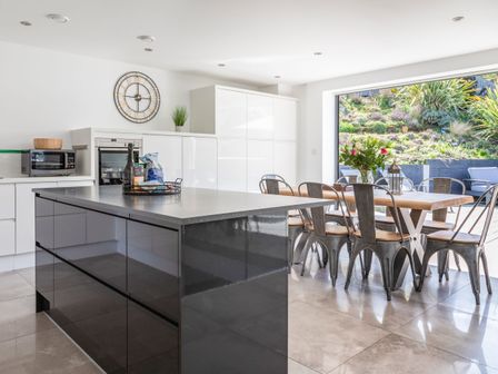 A kitchen with a large island, dining table with metal chairs, and a large open window showing a garden at 7 Beachdown in Challaborough