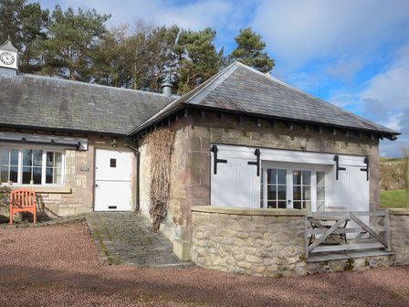 A stone cottage with white doors and windows and a small wooden fence outside Harness Lodge in Seahouses