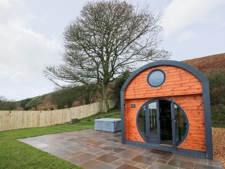 A small wooden pod with circular windows and a glass door on a paved area next to a fenced grassy yard and trees at Callow Pod White Grit near Chirbury
