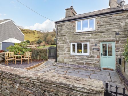 A stone cottage with a green door, two wooden chairs on a patio, and a stone wall at Ael Y Bryn in Ffestiniog