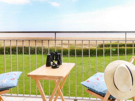 A balcony with a wooden table and chairs with blue cushions overlooking a grassy area and beach at Gull Cottage in Kessingland