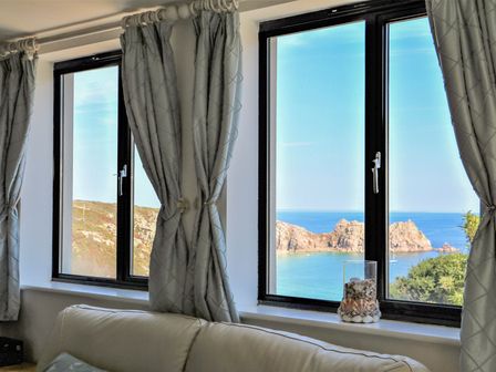 A room with a white sofa and windows showing a rocky beach and blue sea at Beachcomber in Porthcurno near Sennen