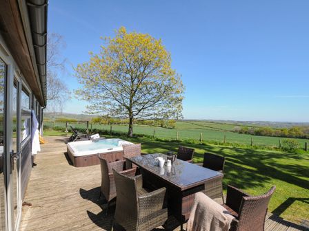 An outdoor wooden deck with a glass table and wicker chairs near a hot tub and a tree with fields in the background at 10 Faraway Fields in Dobwalls