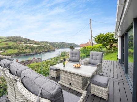 An outdoor seating area with a table and cushioned chairs overlooking a river and green hills at Beach View in Combe Martin